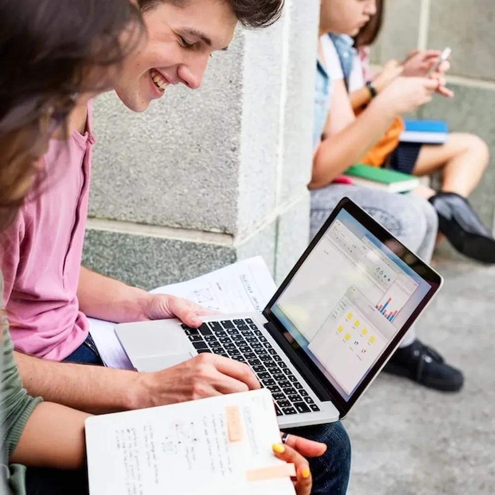 Una estudiante trabaja en una laptop con apuntes mientras sus compañeros estudian cerca