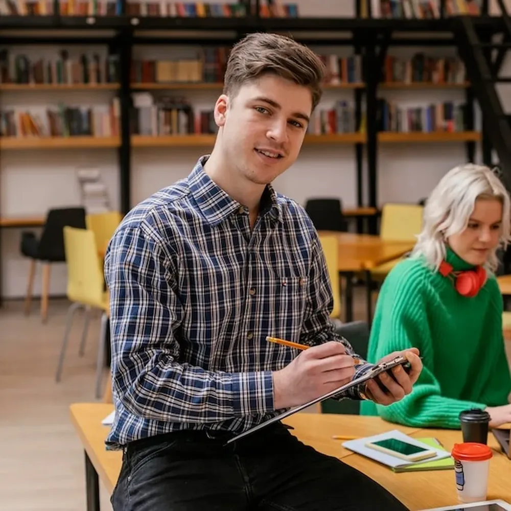 Un estudiante toma notas en una biblioteca universitaria con un compañero detrás